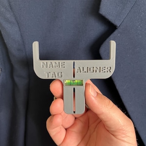 May include: A gray plastic name tag aligner tool with the words "NAME TAG ALIGNER" printed on it. The tool has a level bubble and is held in front of a navy blue blazer.