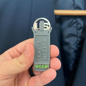 May include: A gray "LAPEL HELPER" tool with a level and "US" emblem. The tool is held in front of a navy blue blazer. The level has a green bubble inside.