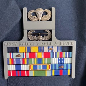 May include: A gray plastic Army badge and rack aligner with various colored ribbons and two metal badges. The text "ARMY BADGE & RACK ALIGNER" is printed on the aligner. The ribbons are red, yellow, blue, and white.