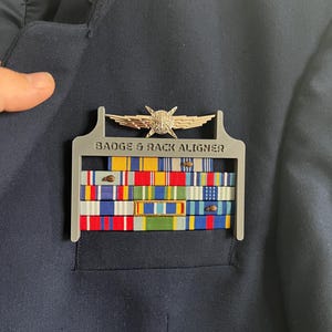 May include: A navy blue jacket with a gray badge and rack aligner holding multiple colorful ribbons. A silver emblem is above the ribbons. The text "BADGE & RACK ALIGNER" is printed on the gray aligner.