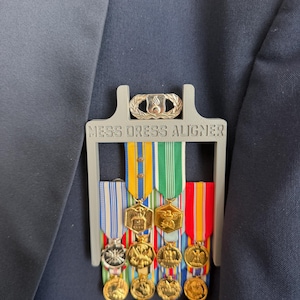 May include: A gray plastic Mess Dress Aligner tool positioned over a dark blue fabric jacket. The tool features a gold-toned metal badge at the top and holds four vertical military ribbon bars, each with multiple gold-toned circular medals attached.