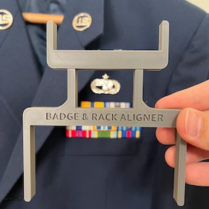 May include: A gray plastic badge and rack aligner tool with the text "BADGE & RACK ALIGNER" is held in front of a navy blue uniform jacket. The jacket has gold-colored buttons and a ribbon rack.