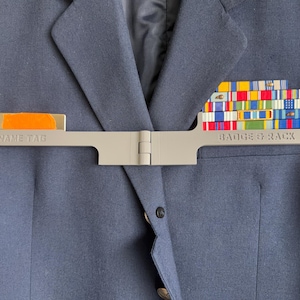 May include: A gray badge and name tag holder attached to a navy blue blazer. The name tag section has an orange rectangle. The badge rack holds several colorful ribbons. The text "NAME TAG" and "BADGE & RACK" are visible.