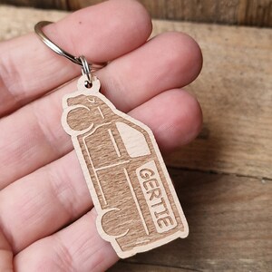 May include: Wooden keychain shaped like a van with the name "Gertie" engraved on the side. The keychain is brown and has a silver metal ring.