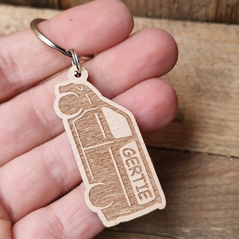 May include: Wooden keychain shaped like a van with the name "Gertie" engraved on the side. The keychain is brown and has a silver metal ring.
