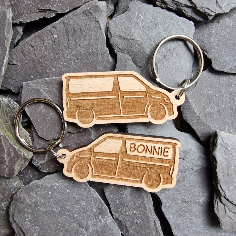 May include: Two wooden keychains shaped like vans. The keychain on the left has a silver key ring. The keychain on the right has the name "BONNIE" engraved on the side.