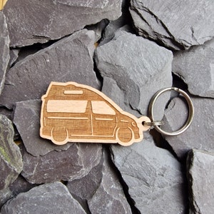 May include: Wooden keychain shaped like a camper van with a silver key ring. The keychain is on a background of gray slate rocks.