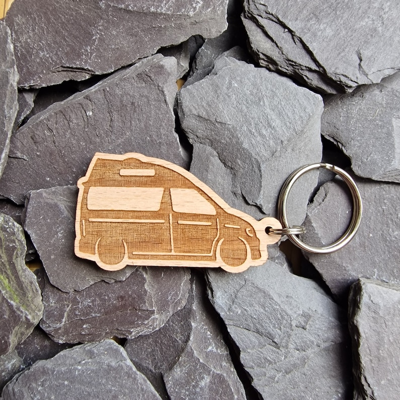 May include: Wooden keychain shaped like a camper van with a silver key ring. The keychain is on a background of gray slate rocks.