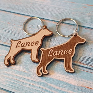 May include: Two wooden dog-shaped keyrings with silver key rings. Both keychains are engraved with the name "Lance". One keyring is light brown and the other is dark brown.
