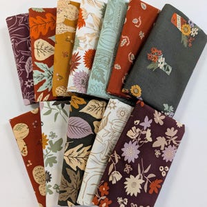 May include: A collection of twelve folded fabric squares featuring autumn-themed patterns. The designs include various illustrations of leaves, pumpkins, flowers, and coffee-related motifs in a color palette of deep red, orange, brown, cream, and sage green.