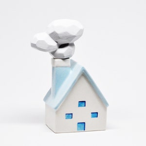 May include: A white ceramic house ornament with a light blue roof and chimney. The house features four blue square windows and a stylized white smoke plume. The ornament is set against a white backdrop.