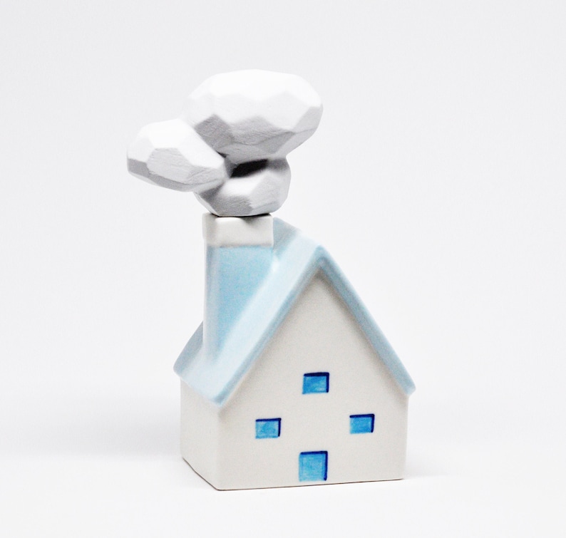 May include: A white ceramic house ornament with a light blue roof and chimney. The house features four blue square windows and a stylized white smoke plume. The ornament is set against a white backdrop.
