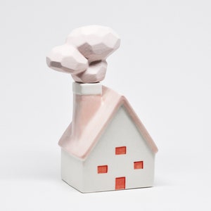 May include: A white ceramic house ornament with a pink roof and chimney. The house features four red square windows and a geometric cloud-shaped chimney topper. The ornament is a decorative item.