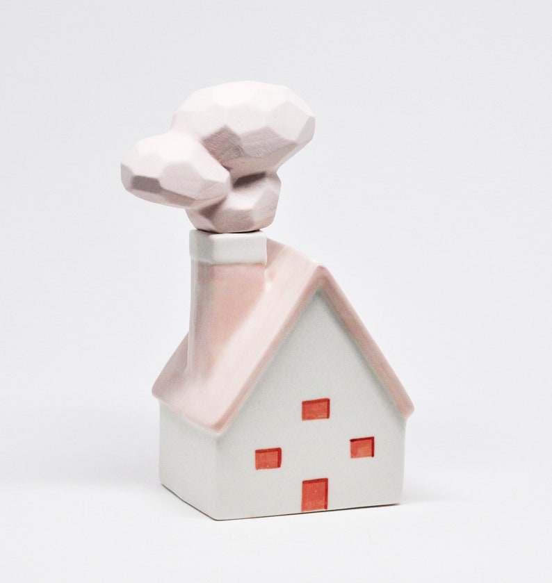 May include: A white ceramic house ornament with a pink roof and chimney. The house features four red square windows and a geometric cloud-shaped chimney topper. The ornament is a decorative item.