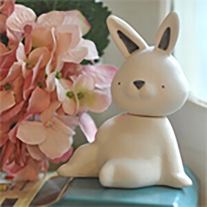 Może przedstawiać: Kremowa ceramiczna figurka królika z czarnymi akcentami na uszach i nosie. Królik siedzi. Różowe kwiaty w tle sugerują wiosenny lub wielkanocny motyw.
