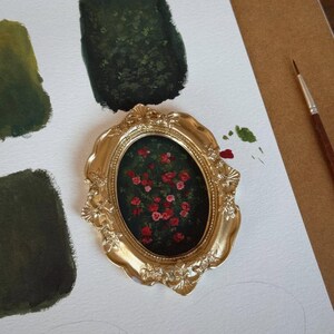 May include: A small, oval painting of red roses in a gold ornate frame. The painting is on a white surface with green paint swatches and a paintbrush. The frame has a detailed, vintage design.