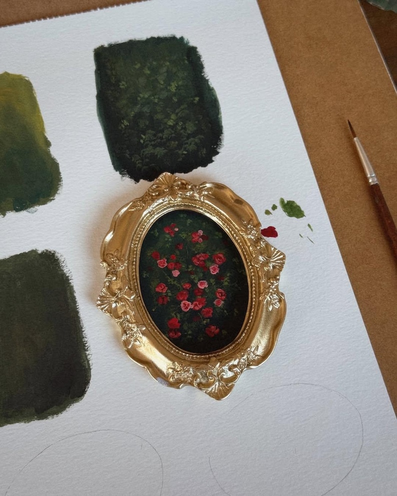 May include: A small, oval painting of red roses in a gold ornate frame. The painting is on a white surface with green paint swatches and a paintbrush. The frame has a detailed, vintage design.