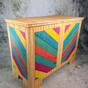 May include: A wooden chest with a light brown finish and colorful diagonal stripes in shades of yellow, blue, green, pink, and red. The chest has a rectangular shape with a flat top and four short legs.