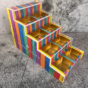 Könnte beinhalten: Mehrstufiger Präsentationsständer mit vertikalem Streifendesign in Regenbogenfarben. Der Ständer hat sechs goldfarbene Metallbehälter. Der Text "CANDY STEPS" ist auf der Seite sichtbar.