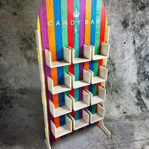 Puede incluir: Un expositor de barra de caramelos de madera multicolor con un fondo a rayas de arcoíris. La pantalla tiene varios estantes pequeños y las palabras "CANDY BAR" están impresas en la parte superior. El expositor es de madera y tiene un gráfico de corona.