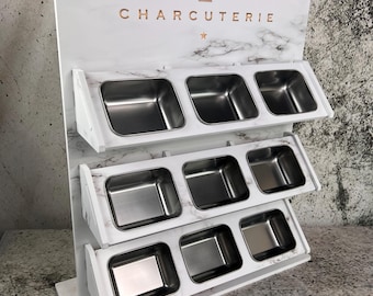 May include: A white charcuterie display stand with a marble pattern and stainless steel containers. The word "CHARCUTERIE" is displayed in gold lettering. The stand has three tiers of containers.
