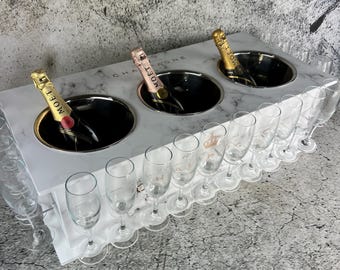 May include: A white marble champagne cooler with three separate compartments for bottles and a built-in rack for champagne flutes. The cooler has a black inscription that reads "Champagne".