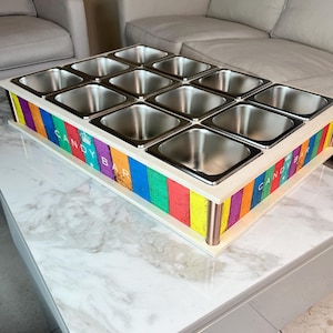 Puede incluir: Una exhibición de barra de dulces con doce recipientes de acero inoxidable. El marco de madera está pintado con rayas verticales de varios colores, incluyendo rojo, amarillo, azul y verde. Las palabras "CANDY BAR" están impresas en el lateral.