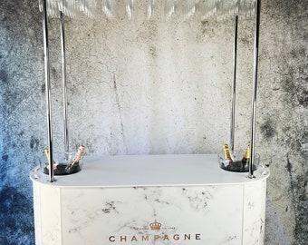 May include: White mobile champagne bar with a marble-patterned surface and a chrome frame. The bar has a top shelf with a row of glass champagne flutes hanging from it. The bar also has two ice buckets with bottles of champagne in them. The bar has a sign that says "FONDEE EN 1743 CHAMPAGNE" with a crown above the word "CHAMPAGNE".