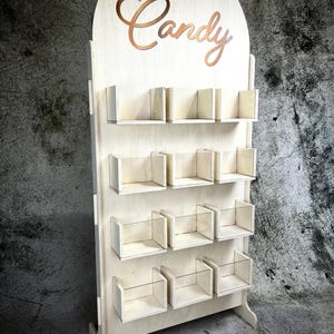 Op de afbeelding: Een houten snoepdisplaystandaard met meerdere planken en kleine kubusachtige compartimenten. Het woord "Candy" is in een cursief lettertype uitgesneden aan de bovenkant van de standaard. De standaard is gemaakt van lichtgekleurd hout.