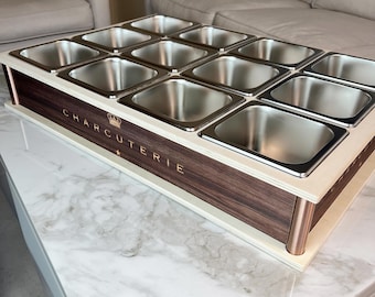 May include: A charcuterie board with twelve stainless steel containers set into a wooden frame. The frame has a dark wood front with the word "CHARCUTERIE" in gold lettering. The board is on a white and marble surface.