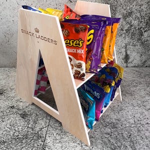 Op de afbeelding: Een lichtgekleurde houten snackdisplaystandaard gevuld met diverse snackzakken. De standaard heeft een driehoekig ontwerp met de tekst "SNACK LADDERS" aan de zijkant. De zakken bevatten Reese's, Nerds en andere kleurrijke snackmerken.