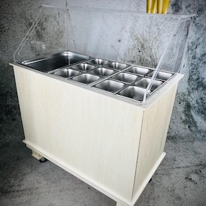 May include: A light-colored wooden food display cart with a clear plastic sneeze guard. The cart has multiple stainless steel containers for food storage and is mounted on wheels for easy mobility. A stack of gold-colored cups sits on top.
