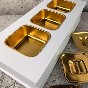 Könnte beinhalten: Ein weißes Serviertablett mit drei goldfarbenen quadratischen Behältern. Zusätzliche goldfarbene quadratische Tabletts sind sichtbar. Die Behälter sind in die weiße Oberfläche eingelassen und erzeugen einen sauberen, modernen Look. Die Tabletts dienen wahrscheinlich zum Servieren von Speisen oder zum Präsentieren von Artikeln.
