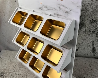 May include: A white charcuterie display stand with a marble pattern and gold-colored containers. The word "CHARCUTERIE" is printed in gold lettering at the top, along with a crown and star symbol. The stand has multiple levels with square containers.
