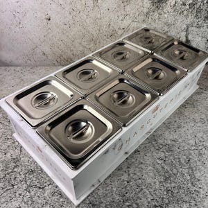 Peut inclure: Un plateau de charcuterie blanc avec un motif marbré, contenant huit récipients en acier inoxydable avec des couvercles. Le mot "CHARCUTERIE" est imprimé sur le côté du plateau.