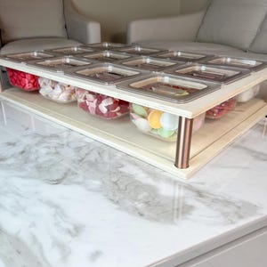Op de afbeelding: Een houten snoepdisplaystandaard met twee niveaus en doorzichtige plastic containers gevuld met kleurrijke snoepjes. De standaard heeft een lichte houten afwerking en koperkleurige steunpoten. De containers zijn afgedekt met roestvrijstalen deksels.