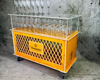 May include: A yellow wooden cart with a clear acrylic top and a diamond-shaped lattice design on the sides. The cart has a sign that says "Champagne" and is on wheels. The cart is filled with champagne glasses.