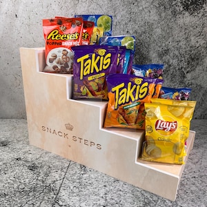 Op de afbeelding: Een houten displaystandaard met meerdere niveaus, gevuld met snackzakken. De snacks zijn onder meer Reese's, Takis Fuego en Lay's Classic chips. De standaard is voorzien van de tekst "SNACK STEPS" in gouden letters.