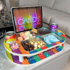 May include: A colorful candy treat bar with a tablet displaying the word "CANDY" in neon lights. The bar features compartments filled with snacks, including popcorn, marshmallows, and cotton candy. A can of soda and a small bowl of pink candies are also visible.