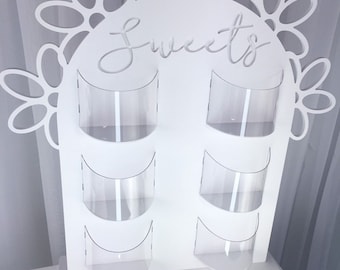 Puede incluir: Un soporte de buffet de dulces de madera blanca con seis recipientes de plástico transparente y un diseño floral en la parte superior. El soporte tiene la palabra "Sweets" escrita en él.