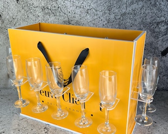 May include: Yellow cardboard box with a black handle and a clear acrylic insert to hold six champagne flutes. The box has the text "Veuve Clicquot Reims France" printed on the front.