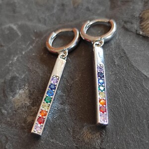 May include: Silver hoop earrings with a rectangular charm, each featuring a rainbow of gemstones. The earrings are displayed on a dark, textured surface. Text: Martina's Klunker's (O275).