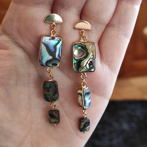 Può includere: Orecchini pendenti dorati con perline rettangolari in madreperla di abalone iridescente. Gli orecchini hanno un semicerchio dorato in alto e una catena con tre perline.