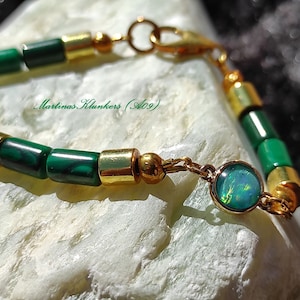 Puede incluir: Un brazalete verde y dorado con un pequeño dije de ópalo. El brazalete está hecho con cuentas de malaquita y oro. El dije es un ópalo redondo con un brillo iridiscente verde, azul y dorado.