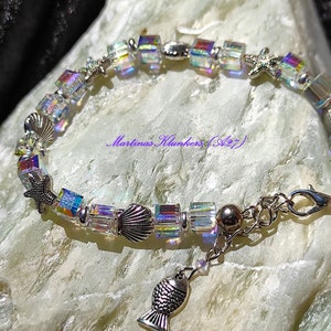 Op de afbeelding: Iridescent kristallen armband met zilverkleurige schelp-, zeester- en visbedels. De armband heeft vierkant geslepen kristallen en een karabijnsluiting. De tekst "Martinas Klunkers (A27)" is zichtbaar.