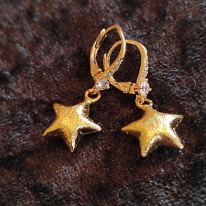 Può includere: Un paio di orecchini a forma di stella color oro. Ogni orecchino presenta una chiusura a leva con piccole pietre trasparenti e un ciondolo a forma di stella pendente. Gli orecchini sono esposti su una superficie strutturata marrone scuro. Il testo "Martina's Klunker's (0298)" è nell'angolo in alto a sinistra.