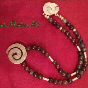 Può includere: Una collana con perline marrone scuro e verdi, con dettagli color oro. La collana presenta due chiusure metalliche color oro, una con un design a spirale e l'altra con un design a incastro. Il testo "Martina's Klunkers K37" è visibile.