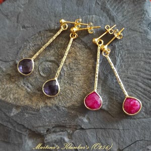 Può includere: Orecchini pendenti dorati con pietre preziose a goccia. Un paio presenta pietre viola scuro, l'altro, rosa vibrante. Gli orecchini hanno steli allungati e strutturati e sono esposti su una superficie grigia strutturata.