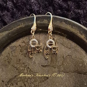 Op de afbeelding: Een paar goudkleurige oorhangers. Elke oorbel heeft een kleine, ronde, met heldere edelstenen bezette halo boven een katten silhouet met een bril. De oorbellen worden gepresenteerd op een donker, getextureerd oppervlak. De tekst "Martina's Klunker's (O 320)" is zichtbaar.