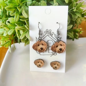 May include: A set of earrings and stud earrings featuring a dog face design. The earrings are brown with a silver hook. The stud earrings are also brown. The earrings are displayed on a white card with a green leafy background.